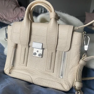 3.1 Phillip Lim Pashli Mini Satchel - Säljer min Philip lim väska i modellen Pashli Mini Satchel🥰 handväskan är krämvit med silverfärgade metalldetaljer och två dragkedjor framtill. Väskan har både handtag och axelrem, samt ett innerfack med dragkedja. Tillverkad i strukturerat skinn. Orginalpriset är 9100kr, skriv till mig vid intresse så kan vi diskutera pris! 