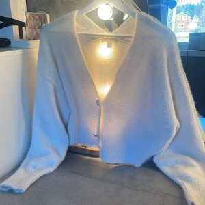 Vit fluffig kofta från H&M med glittriga knappar - Supermjuk och fluffig vit kofta från H&M med v-ringning och två glittriga blomformade knappar framtill. Koftan har långa ärmar och ribbade muddar. Perfekt för lager-på-lager och en mysig look.
