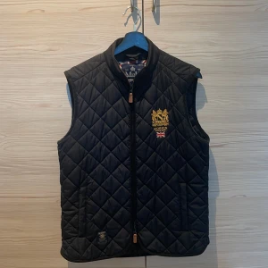Svart quiltad väst från Morris - Stilren svart quiltad väst från Morris med guldbroderad logga. Västen har dubbeldragkedja framtill och klassisk krage. Perfekt för lager-på-lager och snygg till många outfits. Har uppskattat denna väst men har tyvärr växt ur den. Pris kan diskuteras!!!