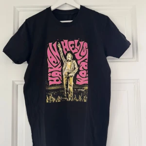 Svart Håkan Hellström t-shirt med tryck - Svart t-shirt med färgglatt tryck av Håkan Hellström på framsidan och turnédatum på baksidan. Trycket är i rosa och gult med retrostil. Perfekt för fans som vill visa sitt stöd för Håkan Hellström Live 2023.
