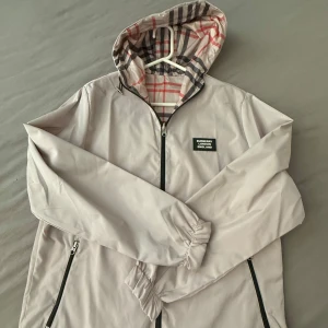 Beige vindjacka från Burberry - Snygg beige vindjacka från Burberry med klassiskt rutigt foder i huvan och svart dragkedja. Jackan har två fickor med dragkedja och en patch med Burberry London England på bröstet. Alla storlekar är sålda förutom storlek S passa på och köp det kan sänka vid en snabb affär.