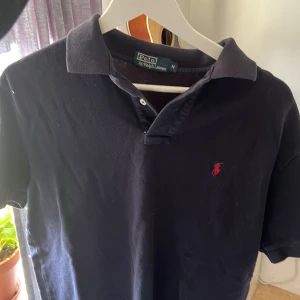 Ralph Lauren pike - Säljer en klassisk mörkblå pikétröja från Ralph Lauren med den ikoniska röda loggan broderad på bröstet. Tröjan har korta ärmar och en stilren krage. Perfekt för en avslappnad men ändå stilfull look. Storlek M använd fåtal gånger🍾