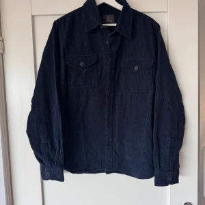 Mörkblå overshirt i manchester från Less Worries - Säljer en mörkblå overshirt i manchestertyg från Less Worries. Skjortan har två bröstfickor med knappar och klassisk krage. Perfekt att bära som lager över en t-shirt eller tröja. Helt oanvänd med prislapp kvar- nypris 1499kr