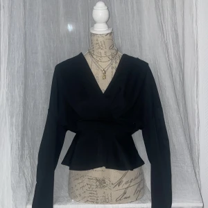 Svart omlottopp med v-ringning - Snygg svart omlottopp med v-ringning och markerad midja. Toppen har långa ärmar och en lätt utsvängd peplum nedtill. Perfekt för att skapa en stilren och trendig look. 