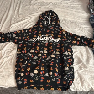 Svart hoodie med svampmönster från SQRTN Company - Säljer en svart hoodie från SQRTN Company med färgglatt svampmönster och vit text 'Norrland' på bröstet. Tröjan har huva med vita snören och en stor magficka. Perfekt för dig som gillar unika prints och streetwear.