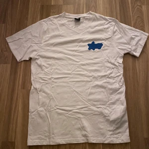 Vit t-shirt med blå broderad detalj - Vit t-shirt från FSBN och en blå broderad detalj på bröstet. Klassisk passform och korta ärmar, perfekt för en enkel och stilren look.