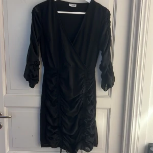 Svart omlottklänning med rynkade detaljer - Säljer en svart omlottklänning med trekvartsärm och rynkade detaljer längs sidorna. Klänningen har v-ringning och är tillverkad i ett lätt, glansigt material. Går att justera längre/kortare 