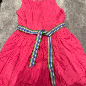 Klänning - Säljer en rosa klänning från Polo Ralph Lauren med ärmlös design och rund hals. Klänningen har ett färgglatt randigt bälte i midjan och knappar längs ryggen. Perfekt för sommarens tillfällen! 154 cm. 