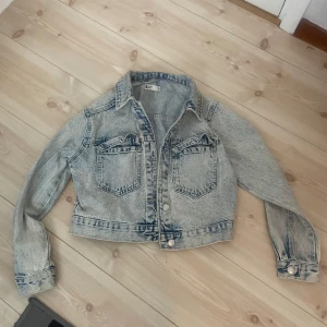 Ljusblå croppad jeansjacka från Gina  - Säljer en ljusblå croppad jeansjacka i storlek XS med klassisk krage och två bröstfickor med lock. Jackan har långa ärmar och knappar framtill samt vid ärmsluten. Perfekt för en avslappnad och trendig look. 