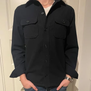 Svart overshirt från Riley - Stilren marinblå overshirt från Riley med klassisk krage och två bröstfickor med lock. Skjortan har långa ärmar och knäppning framtill. Perfekt att bära över en t-shirt för en avslappnad look. Nyskick använd några få gånger. Storlek M. Nypris ligger på runt 1200 men mitt pris är 549! Sjukt bra pris 🙌