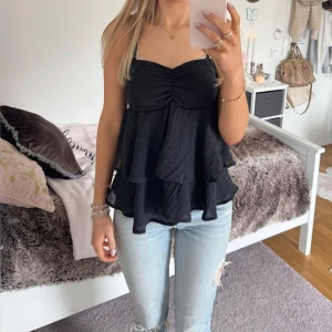 Svart volangig linnetopp - Säljer en svart linnetopp med smockad överdel och flera lager volanger. Toppen har tunna axelband och en lätt rynkad detalj framtill. Perfekt att styla med jeans eller kjol för en trendig look. ⊹ jag är 173cm & stl S/36/28 för referens ⊹ läs POLICY (i gillade inlägg) innan köp ⊹ använd gärna ”Köp” ⊹ 