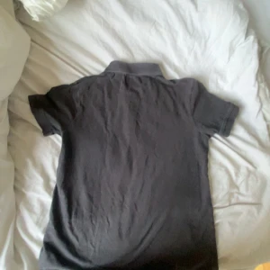 Svart pikétröja från Lyle & Scott - Säljer en klassisk svart pikétröja från Lyle & Scott med gul broderad logga på bröstet. Tröjan har korta ärmar, krage och knappar framtill. Perfekt för en stilren och avslappnad look.