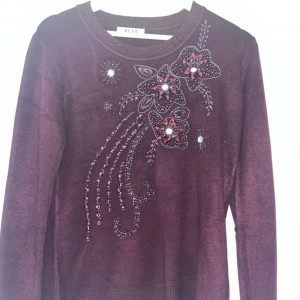 Vinröd tröja med blommor och pärlor från Q.L.F.X - Mörkröd tröja med långa ärmar och rund halsringning. Framsidan är dekorerad med blommor, pärlor och glittriga detaljer. Perfekt för dig som vill sticka ut lite extra med en unik design.