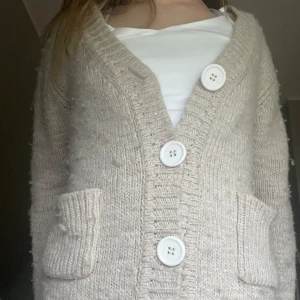 Beige stickad kofta från Gul&Blå - Mysig beige stickad kofta från Gul&Blå med stora vita knappar och två fickor framtill. Koftan har lång ärm och en klassisk v-ringad design, perfekt att slänga på sig över en t-shirt eller topp.