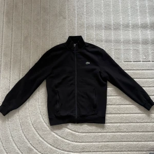 Svart zip-jacka från Lacoste - Säljer en svart zip hoodie från Lacoste med hög krage. Jackan har två fickor framtill och den klassiska Lacoste-loggan på bröstet. • Storlek M • Frakt tillkommer  • Har du några frågor är det bara att skicka ett meddelande, så svarar jag så fort jag kan.☺️ Ha en fin dag!💕♻️