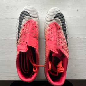 Nike fotbollsskor med snörning - Säljer ett par Nike fotbollsskor i röd och vit färg med svart swoosh. Skorna har snörning och är märkta med ACC (All Conditions Control) för extra grepp i alla väder. Perfekta för dig som vill ha snygga och funktionella skor på planen.