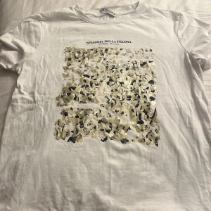 Vit t-shirt med leopardmönster från Zara - PRISET GÅR ATT DISKUTERA!!!          Vit t-shirt från Zara med ett stort glittrigt leopardmönster i guld och svart framtill. Trycket har texten 'SPIAGGIA DELLA PELOSA' ovanför. Klassisk passform och korta ärmar, tillverkad i mjuk bomull. Tyvärr så har lappen där uppe gått av på ena sidan.