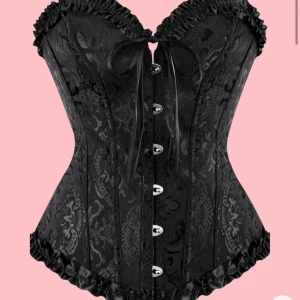 Svart korsett med spetsdetaljer - Säljer en svart korsett med fina spetsdetaljer och snörning framtill. Modellen har en klassisk form och dekorativa volanger längs kanterna. 