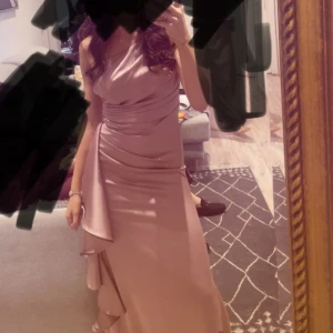 Rosa  långklänning med volangdetaljer - Elegant långklänning i rosafärgat  satin med omlott och volangdetaljer längs kjolen. Klänningen har en axelbandslös design och markerad midja för en stilren siluett. Perfekt för bal eller festliga tillfällen.