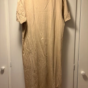 Beige långklänning från 157 - Enkel och stilren beige långklänning från 157 i mjuk bomull. Klänningen har korta ärmar och rak passform, perfekt för en avslappnad look. Materialet är 100% bomull och färgen är neutral beige.