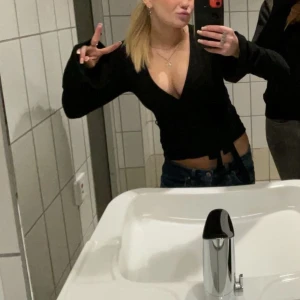 Svart omlottopp med knyt - Säljer en svart omlottopp med knyt i midjan. Toppen har långa ärmar och djup v-ringning, perfekt att styla över ett linne eller topp. Enkel och stilren design som passar till mycket.