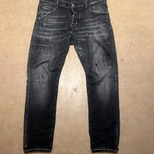 Svarta jeansbyxor från Dsquared2 - Säljer ett par svarta jeansbyxor från Dsquared2 i storlek 50. Jeansen har slitna detaljer, diskreta hål och patchar framtill samt broderad DS2-logga på bakfickan. Klassisk femficksmodell med knappgylf och raka ben.