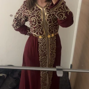 Kaftan marrockansk  - Säljer en vinröd långklänning med vackra guldfärgade broderier och dekorativa knappar framtill. Klänningen har långa ärmar och en elegant, traditionell look med markerad midja. Perfekt för speciella tillfällen.