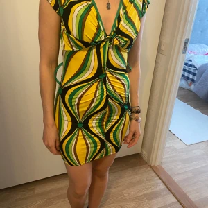 Färgglad kortklänning vintage  - Säljer en kort klänning med färgstarkt retroinspirerat mönster i gult, grönt, vitt och svart. Djup v-ringning och rynkade detaljer framtill. Vintage.Klänningen har korta ärmar och figurnära passform. Perfekt för dig som vill sticka ut!stretch passar 34-38
