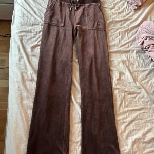 Bruna velourbyxor från Juicy Couture - Mysiga bruna velourbyxor från Juicy Couture med bred resår i midjan, stora framfickor och bakfickor med knappar. Byxorna har broderad JC-logga framtill och vida ben. Perfekta för en avslappnad och trendig look. Bara använda ett fåtal gånger💗