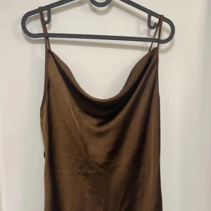 Brun satin slipklänning från Motel - Säljer en brun satin slipklänning från Motel med tunna axelband och snygg draperad urringning. Klänningen har en mjuk glans och är perfekt för dig som gillar minimalistisk stil. Snörre kommer också med för att kunna knyta där bak i midjan!! Passar lika bra till sneakers som klackar.