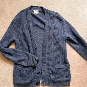 Blå kofta från Woolrich - Mysig blå kofta från Woolrich med knappar framtill och två fickor. Koftan har ribbade muddar och en klassisk v-ringad öppning. Perfekt för lager-på-lager och enkel att matcha med olika outfits.