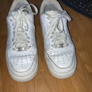 Vita Nike Air Force 1 sneakers - Klassiska vita Nike Air Force 1 sneakers med rund tå och snörning. Skorna har en platt sula och är tillverkade i skinn med diskreta detaljer. Perfekta för en stilren och sportig look.