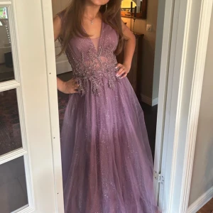 Ljuslila långklänning med silverglitter och broderier - Säljer en ljuslila långklänning med djup v-ringning och glittrigt tyg. Klänningen har vackra broderade detaljer och är ärmlös. Perfekt för bal eller festliga tillfällen. Tyllkjol ger extra volym och en riktigt drömmig känsla. Stl.34/36 och passar för längd 160-170 då längd är uppsydd och kan släppas ut. Använd 1 gång och har inga skador/fläckar. Köpt på FESTIY i Kista galleria. Köpt för 2600.