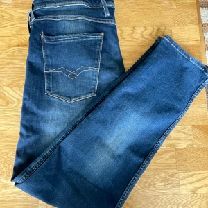 Replay Anbass jeans i blå denim - Säljer ett par blå Replay Anbass jeans med klassisk femficksdesign och snygga kontrastsömmar. Jeansen har normal passform och raka ben, tillverkade i slitstark denim. Perfekta för dig som gillar stilrena och tidlösa jeans. Jeansen har ett litet slitage vid gylfen men det syns ej och är inget problem då de inte är öppet vid gylfen. Vid fler funderingar är det bara att skriva