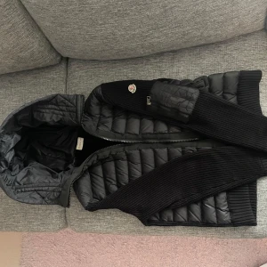 Moncler cardigan Svart dunväst med stickade detaljer från Dunst - Snygg svart dunväst med stickade partier på sidorna och huva. Västen har dragkedja framtill och en klassisk quiltad look. Perfekt för lager-på-lager och passar till många stilar.