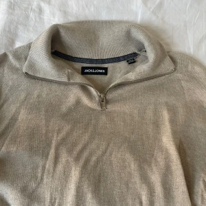 Beige stickad tröja med dragkedja från Jack & Jones - Säljer en beige stickad tröja från Jack & Jones med halv dragkedja vid kragen. Tröjan har lång ärm och ribbade muddar vid ärmslut och nederkant. Perfekt för lager-på-lager under kyligare dagar.