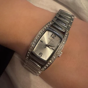 Silver klocka  - Säljer en snygg silverfärgad klocka från Moretime med rektangulär urtavla och glittriga stenar längs kanterna. Klockan har en metallänk och stilren design med siffrorna 12 och 6 markerade på urtavlan.