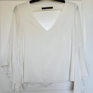 Vit blus med volangärmar från Zara - Säljer en elegant vit blus från Zara Trafaluc Collection med v-ringning och draperad öppen rygg. Blusen har långa, vida volangärmar som ger en snygg och trendig look. Perfekt för dig som vill sticka ut med stilrena detaljer.