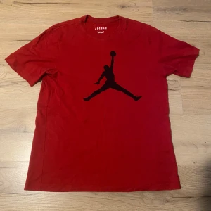 Jordan T-Shirt - Säljer en röd t-shirt från Jordan med den klassiska svarta Jumpman-loggan på bröstet. T-shirten har normal passform och är tillverkad i mjuk bomull. Perfekt för dig som gillar sportig stil och streetwear.