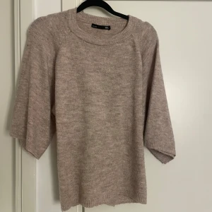 Beige stickad tröja från 157 - Mysig beige stickad tröja från 157 med trekvartsärm och bred ribbad kant i halsen. Baksidan har en snygg detalj med ribbad knytning i midjan som ger en unik look. Perfekt för lager-på-lager under kyliga dagar.