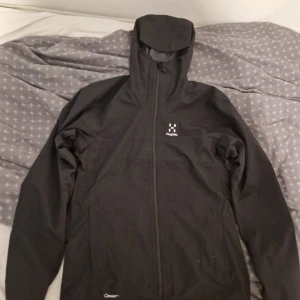 Svart vindjacka från Haglöfs - Säljer en svart vindjacka från Haglöfs med huva och dragkedja framtill. Jackan har minimalistisk design med broderad logga på bröstet och är perfekt för utomhusaktiviteter. Snygg och funktionell jacka som passar till många tillfällen.
