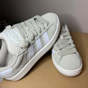 Adidas Campus 00s W ljusgrå/begie sneakers - Säljer ett par Adidas Campus 00s W sneakers i ljusgrå mocka med vita detaljer och klassiska tre ränder på sidorna. Skorna har breda snören och Adidas-logga på plösen. Perfekta för en stilren vardagslook. Aldeig använda prislapp på 
