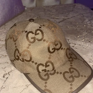 Beige keps från Gucci med GG-mönster - Säljer en beige keps från Gucci med det klassiska GG-mönstret i brunt. Kepsen har böjd skärm och justerbart spänne bak i brunt läder. Tillverkad i bomullsmix och har en exklusiv känsla. Perfekt för dig som gillar lyxiga accessoarer.