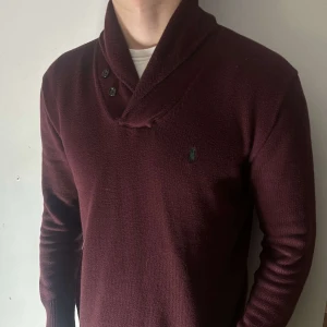 Vinröd stickad tröja med krage från Polo Ralph Lauren - Säljer en vinröd stickad tröja från Polo Ralph Lauren med snygg sjalkrage och tre knappar vid halsen. Tröjan har lång ärm och diskret logga på bröstet. Perfekt för en stilren och bekväm look.