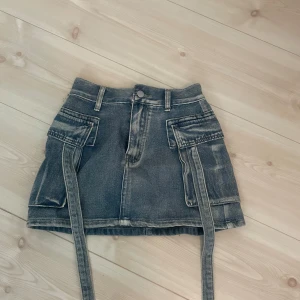 Grå jeanskjol med fickor och bältdetaljer - Säljer en cool grå jeanskjol med stora fickor framtill och bak samt långa bältdetaljer som hänger ner. Kjolen har en kort passform och markerade sömmar för en trendig look. Perfekt för dig som gillar streetstyle.