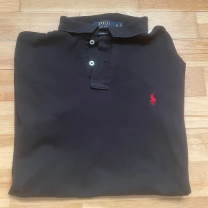 Stilren Svart Ralph Lauren piké - M - Stilren svart Ralph Lauren piké – M  Riktigt snygg och klassisk pikétröja från Ralph Lauren i svart – en sån där garderobsfavorit som alltid funkar. Gjord i 100 % bomull, vilket gör den både mjuk och skön att ha på sig. Den ikoniska loggan sitter på bröstet, och modellen har den klassiska kragen med knappar.  🖤 Färg: Svart 📏 Storlek: M (Byst ca 43 cm / Längd ca 70 cm) 👌 Skick: Mycket bra – inga fläckar, hål eller synligt slitage 👕 Material: Bomull – mjuk, sval och perfekt till våren/sommaren