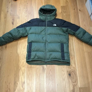 Grön dunjacka från The North Face - Säljer en grön och svart dunjacka från The North Face med huva och dragkedja. Jackan har en pufferstil och två svarta fickor framtill. Perfekt för kyliga dagar och riktigt skön att bära. Storlek XL men passar bättre för L eller M. Köpt för 2500+ men säljer för 1000