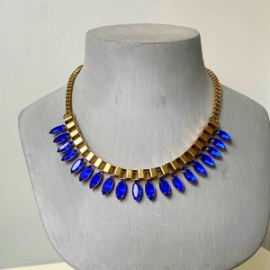 Guldfärgat halsband med blå stenar - Säljer ett snyggt halsband i guldfärgad metall med stora, droppformade blå stenar. Halsbandet har en kedja med justerbar längd och ger en riktigt cool statement-look. Perfekt för dig som vill sticka ut med färg och stil.
