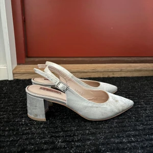 Vita  pumps med klack och dekorerat spänne - Säljer ett par eleganta beige pumps med spetsig tå och bred klack. Skorna har öppen häl och ett dekorativt spänne med pärlor på remmen. .