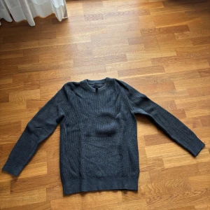 Mörkgrå stickad tröja - Mysig mörkgrå stickad tröja med rund hals och långa ärmar. Perfekt för kyligare dagar och enkel att matcha med olika outfits. Klassisk modell med ribbade muddar vid ärmslut och nederkant.